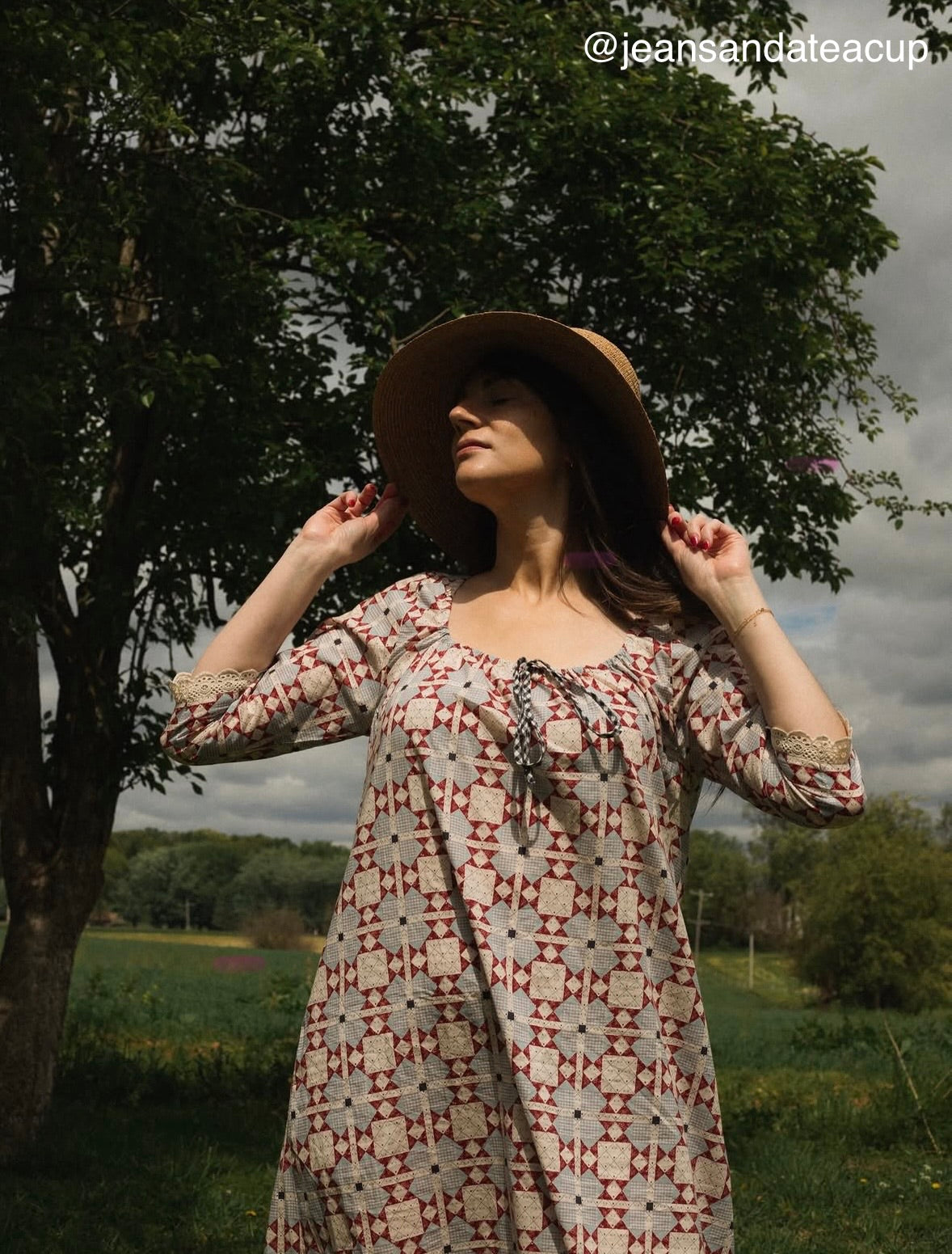 Woman in a patterned dress and hat standing outdoors with trees and sky in the background