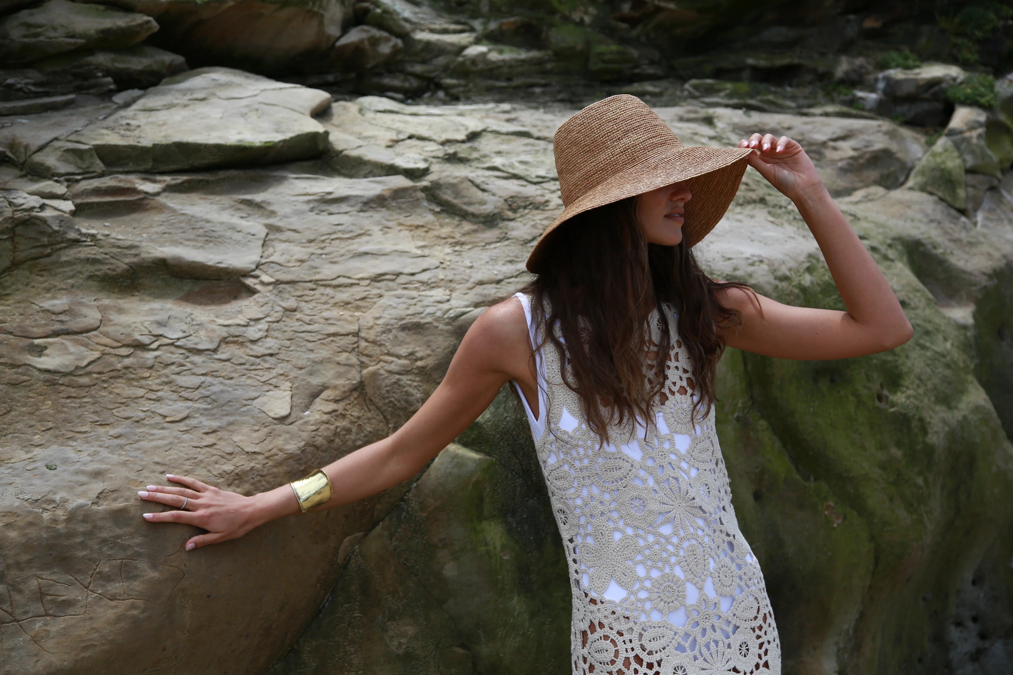 Woman in a white dress and straw hat standing against a rocky wall.