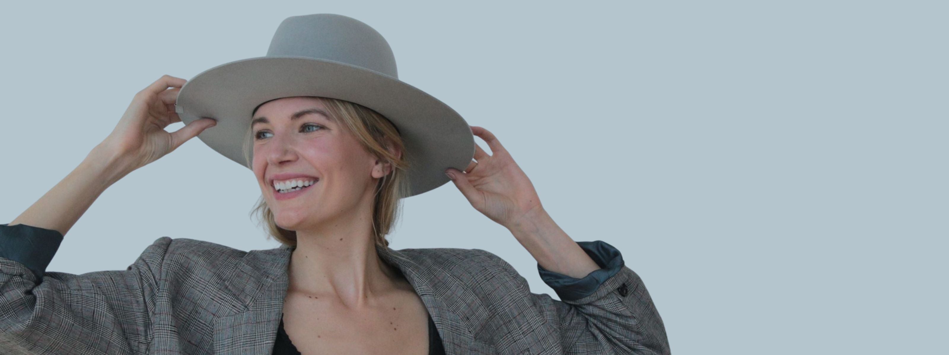 Smiling woman adjusting a wide-brimmed hat, wearing a plaid blazer against a light background.